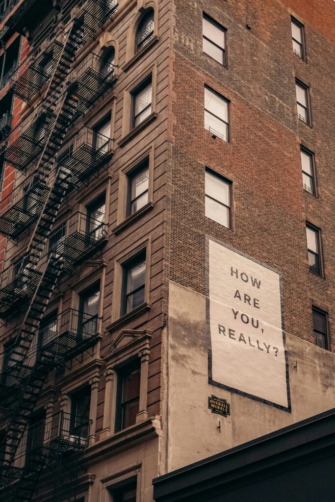 Die Außenwand eines alten, hohen Wohngebäudes aus rotem Klinker. Die triste Erscheinung mit den alten Feuerleitern an der Fassade wird gebrochen durch das großflächig aufgemalte Schild: How are you really?